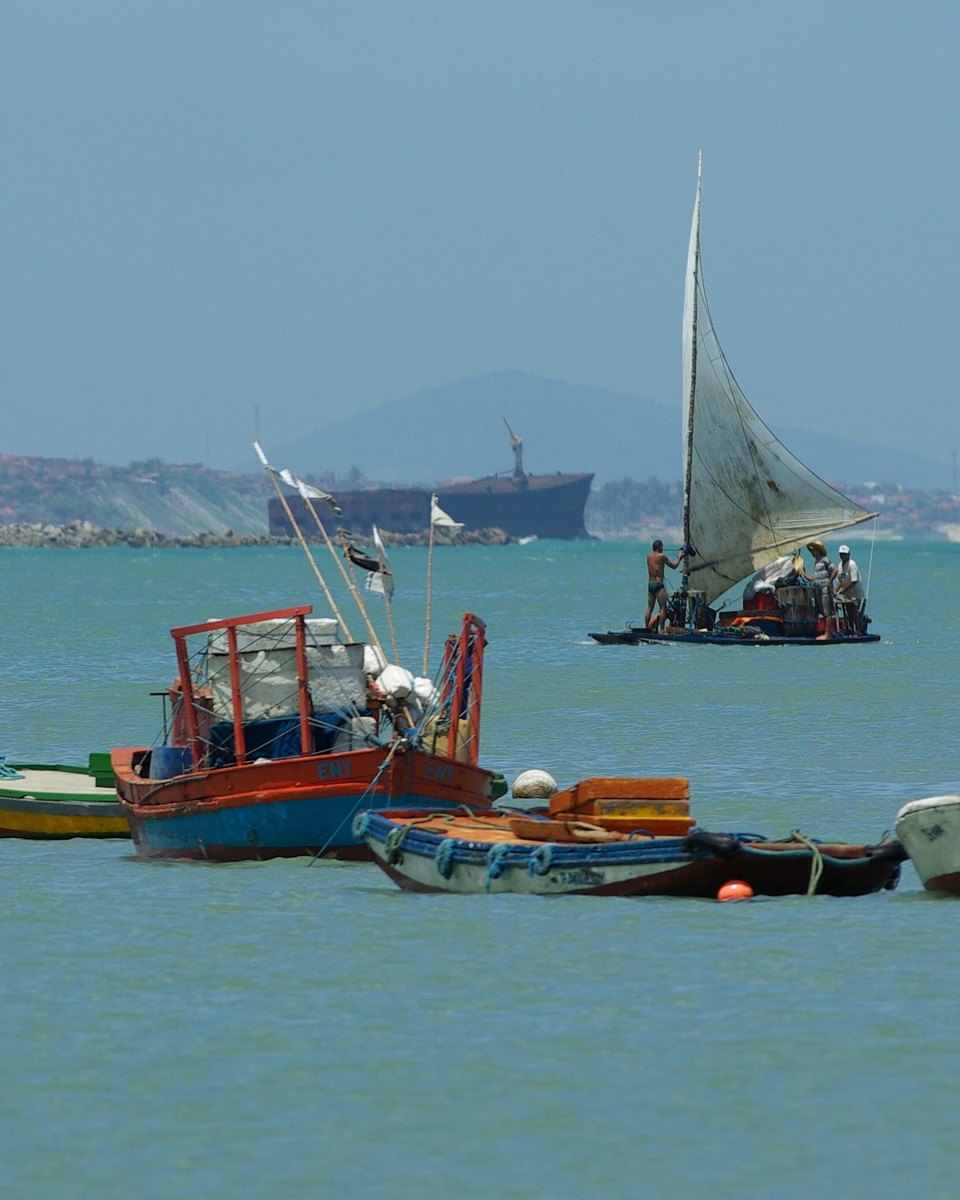 Barcos de pesca coloridos ancorados na enseada do Mucuripe, em Fortaleza, com uma jangada de vela branca navegando ao lado, a água verde e a costa com um navio ao fundo.