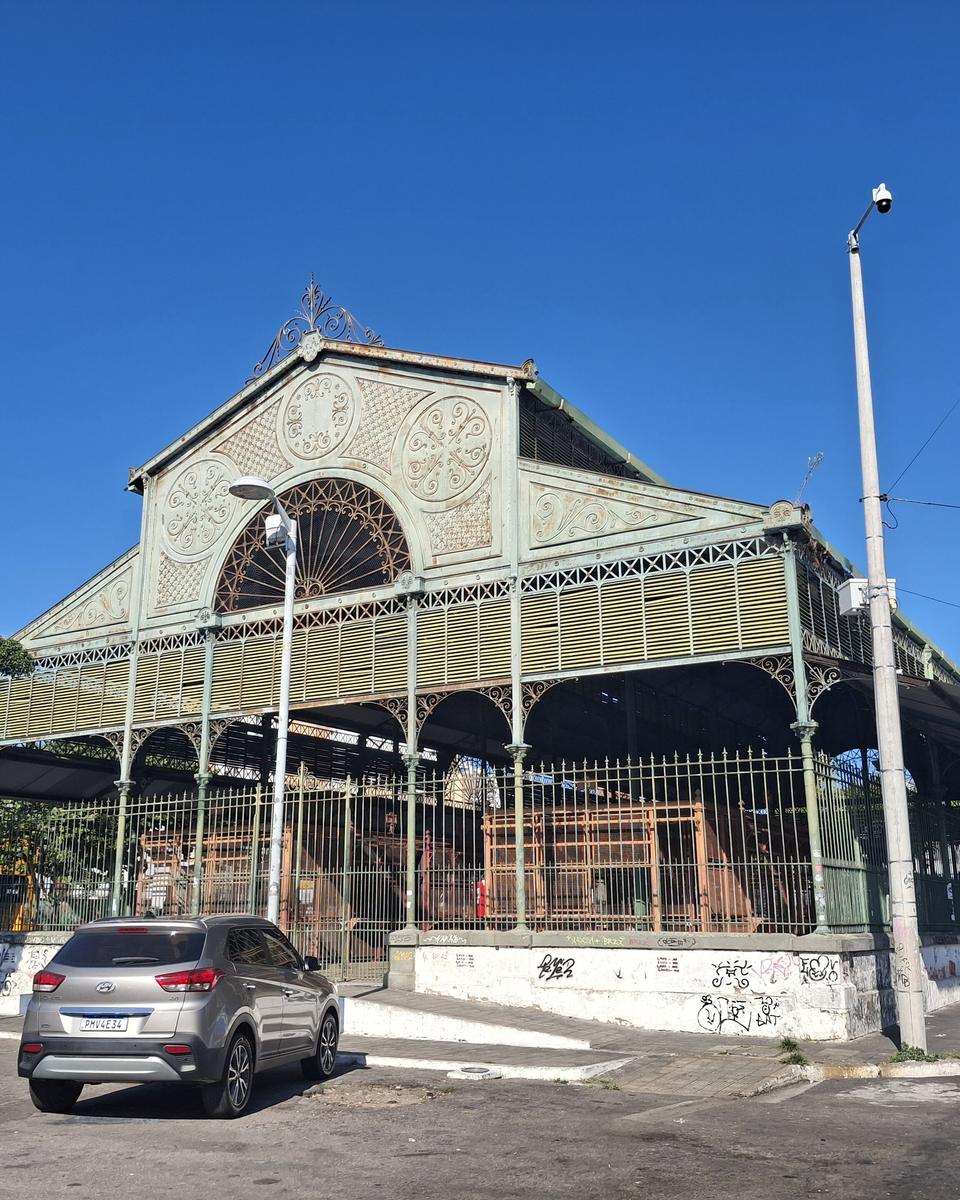 Mercado dos Pinhões, no Centro de Fortaleza, com a estrutura de ferro verde e a fachada em arco rendado, postes de luz e um carro estacionado em frente sob céu azul.