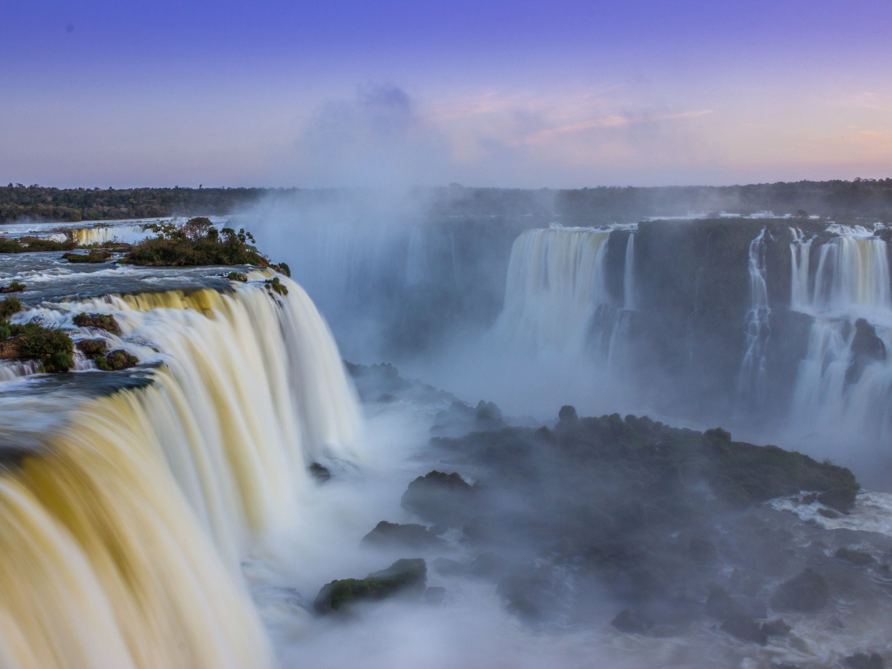 Garganta do Diabo das Cataratas do Iguaçu vista do lado brasileiro ao entardecer, com a água caindo em véu, a névoa subindo e o céu lilás e rosado.