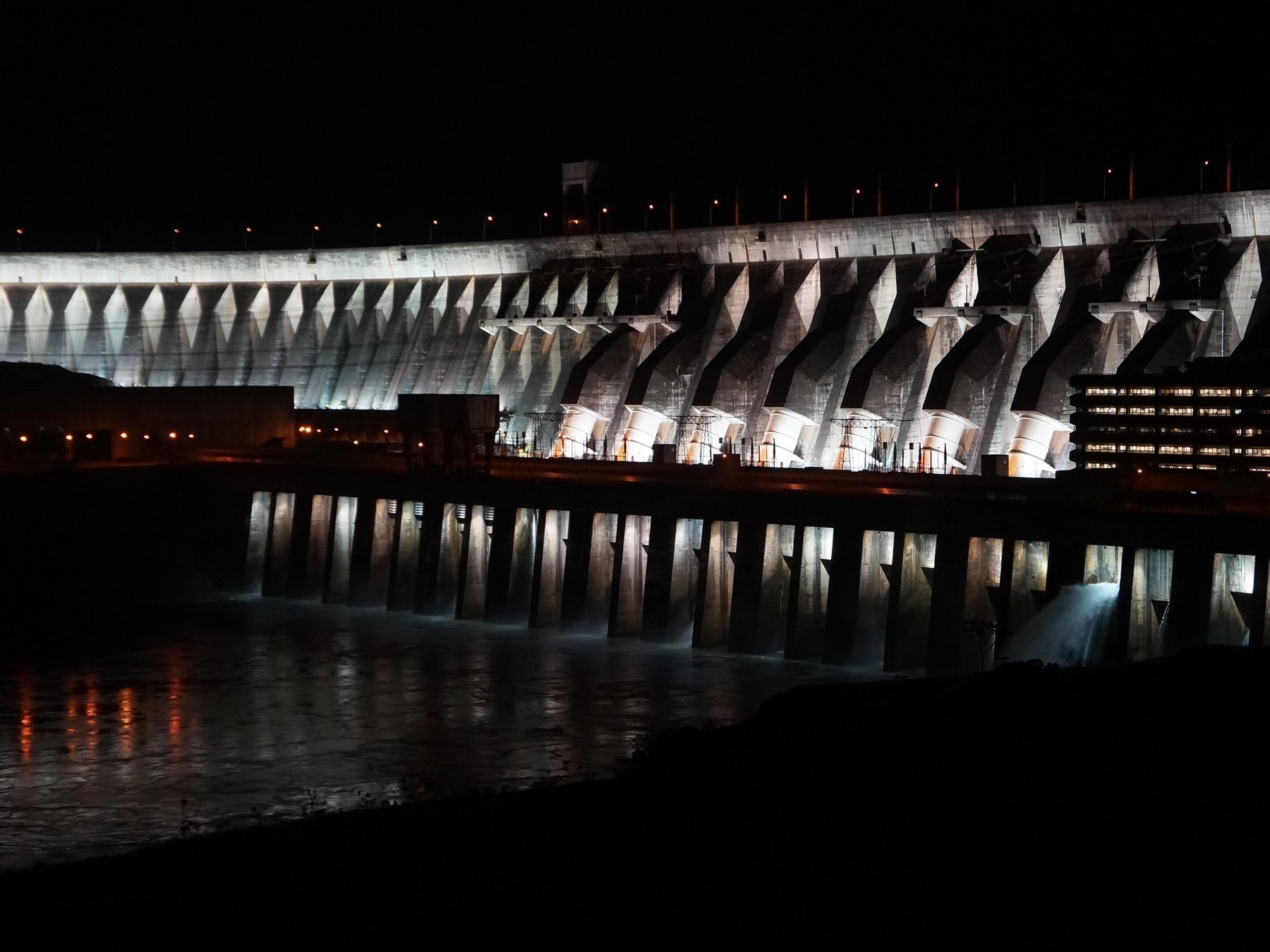Barragem de Itaipu iluminada à noite, com os pilares de concreto e os tubos brancos refletidos no rio Paraná.