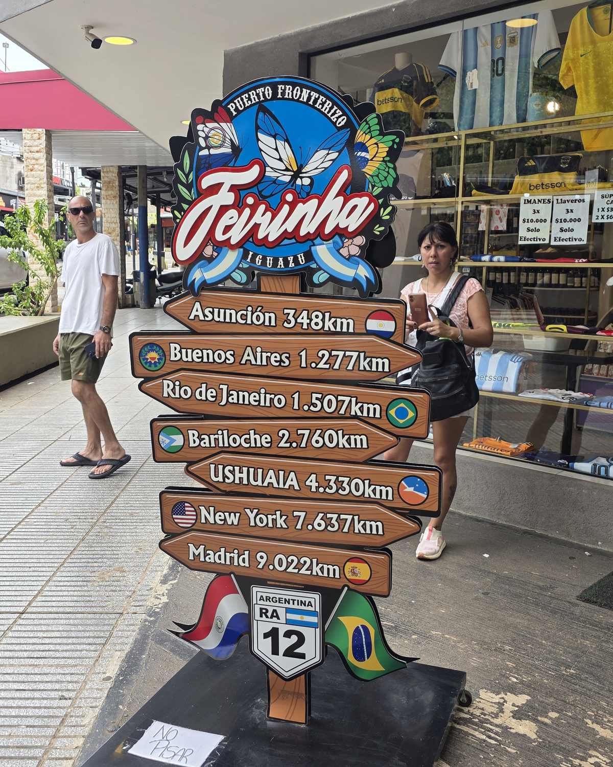 Placa colorida da Feirinha de Puerto Iguazú, com setas de distância para Asunción, Buenos Aires, Rio de Janeiro e outras cidades, e dois turistas passando pela calçada em frente às lojas.