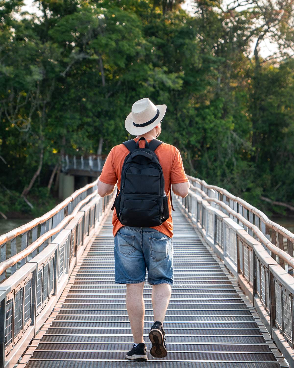 Turista de chapéu, camiseta laranja e mochila caminhando de costas numa passarela de metal sobre o rio Iguaçu, entre a mata, no fim da tarde.