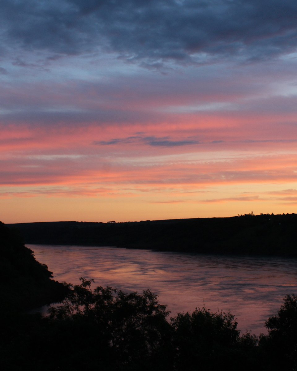 Pôr do sol alaranjado sobre o encontro dos rios Iguaçu e Paraná, visto do Marco das Três Fronteiras, com a mata escura das margens em silhueta.
