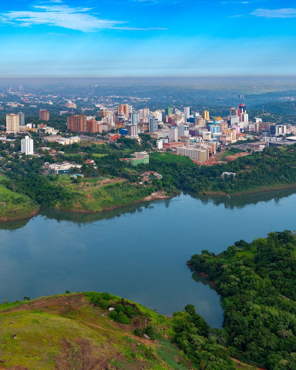 Vista aérea do rio Paraná com a Ponte da Amizade à direita e os prédios de Ciudad del Este, no Paraguai, na outra margem, sob céu azul.