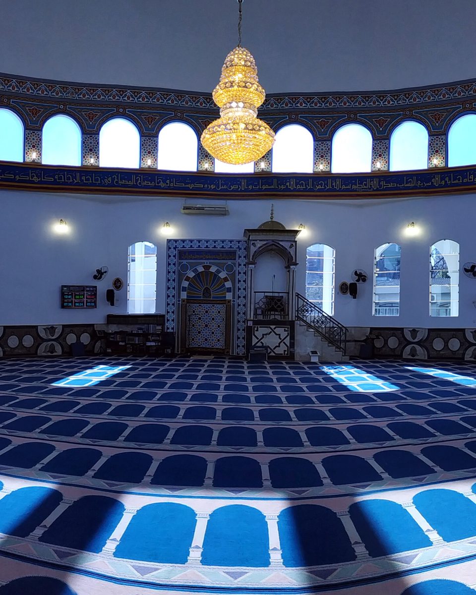 Salão de oração da Mesquita Omar Ibn Al-Khattab, em Foz do Iguaçu, com tapete azul, janelas em arco em volta, um lustre dourado no centro e o nicho de oração decorado ao fundo.