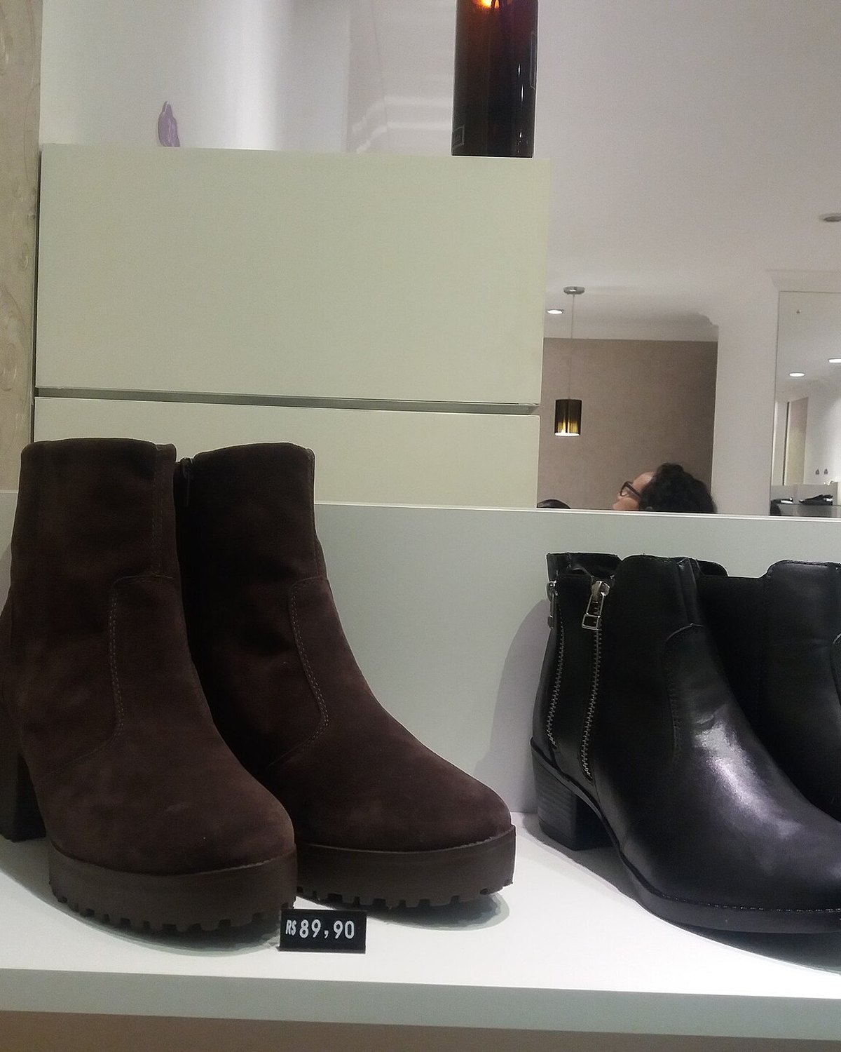 Vitrine de uma loja no Shopping do Calçado de Franca, com uma bota de camurça marrom e uma bota de couro preto na prateleira, cada uma com a etiqueta de preço.