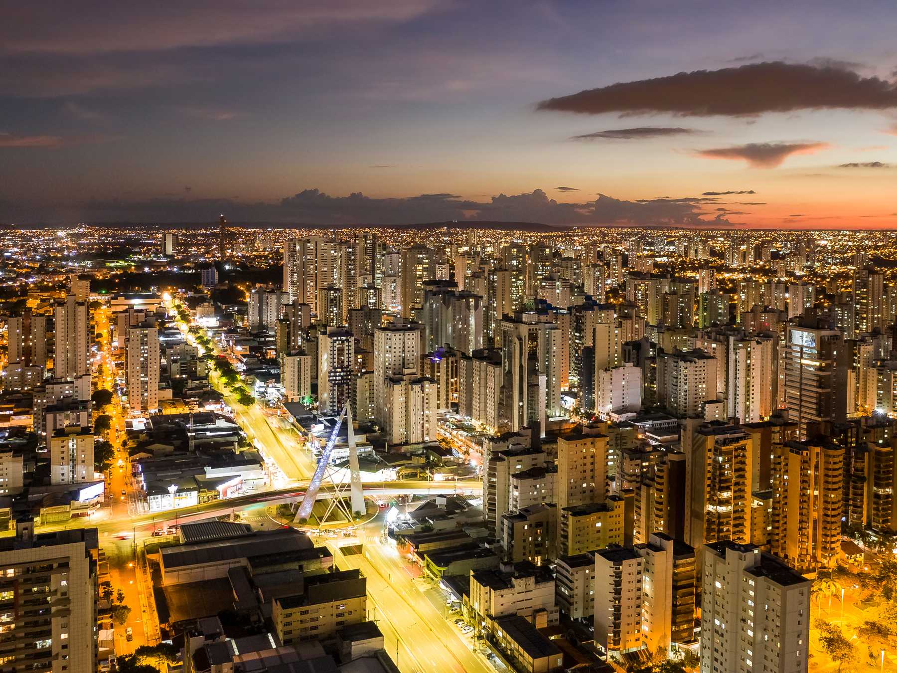 Vista aérea de Goiânia ao anoitecer, com os prédios altos acesos, as avenidas riscadas de luz amarela e o monumento de aço do Viaduto João Alves de Queiroz, na Avenida T-63, sob um céu alaranjado.