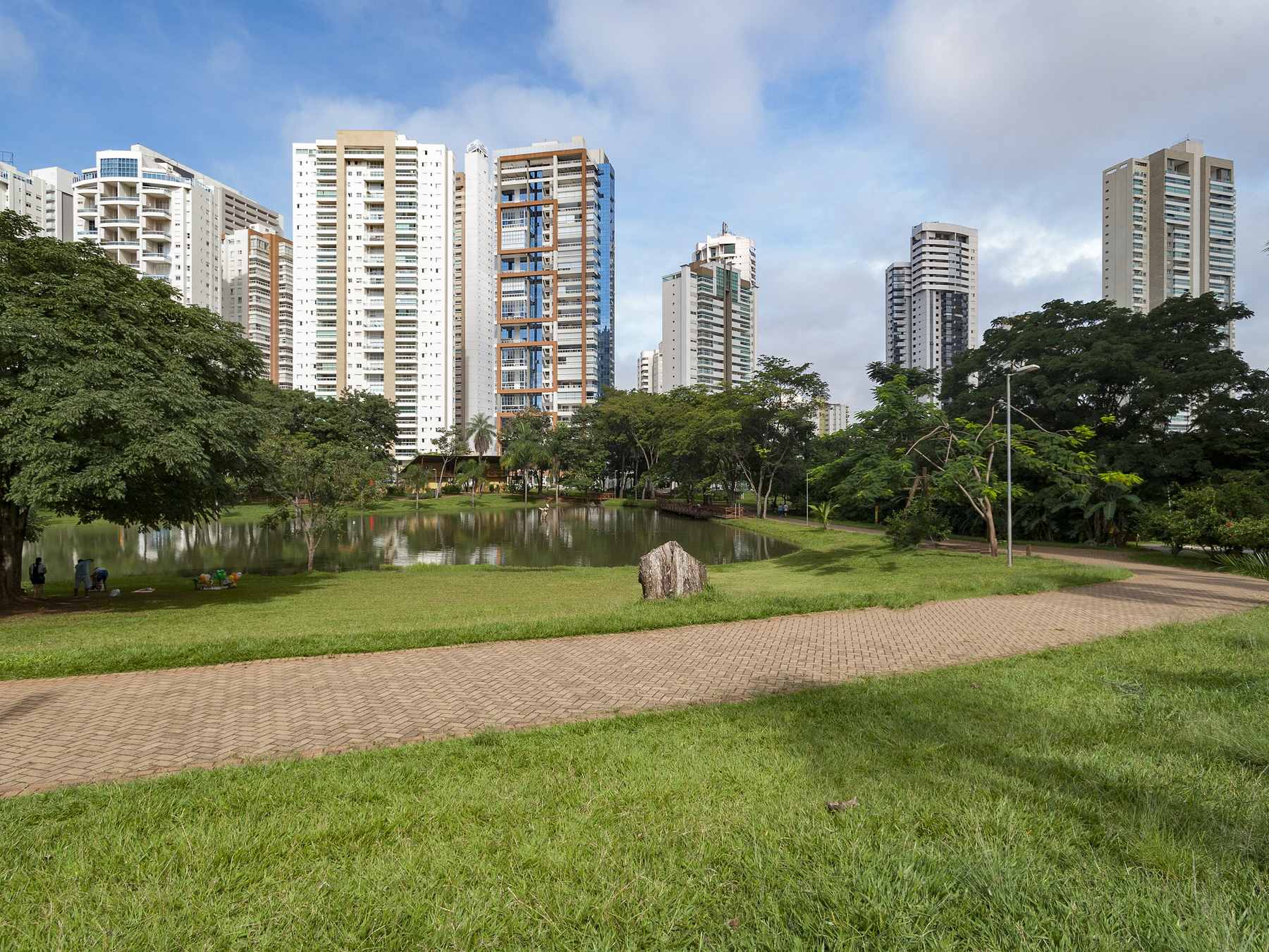 Parque Flamboyant, no Jardim Goiás, com o gramado e a pista de caminhada contornando o lago, árvores grandes fazendo sombra e os prédios altos do bairro ao fundo sob céu azul com nuvens.