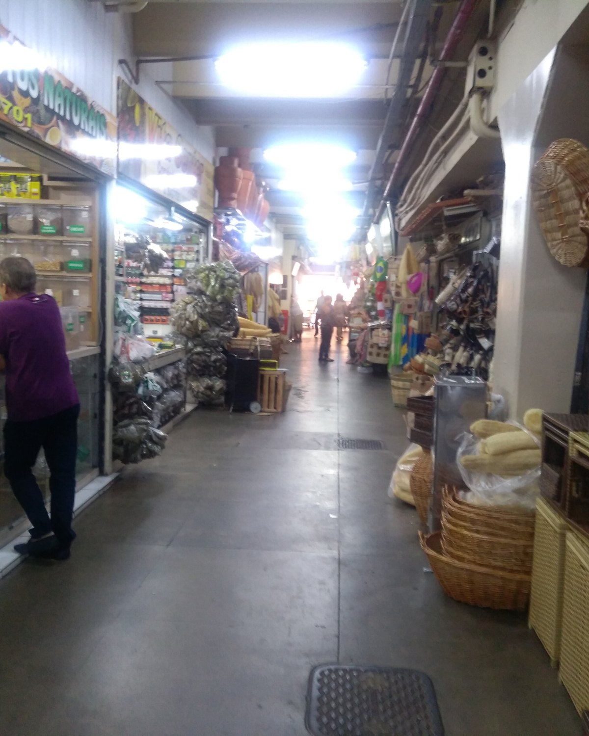 Corredor do Mercado Central de Goiânia com bancas de temperos e produtos naturais de um lado, cestos de palha e chapéus do outro, e um cliente conversando com o comerciante no balcão.