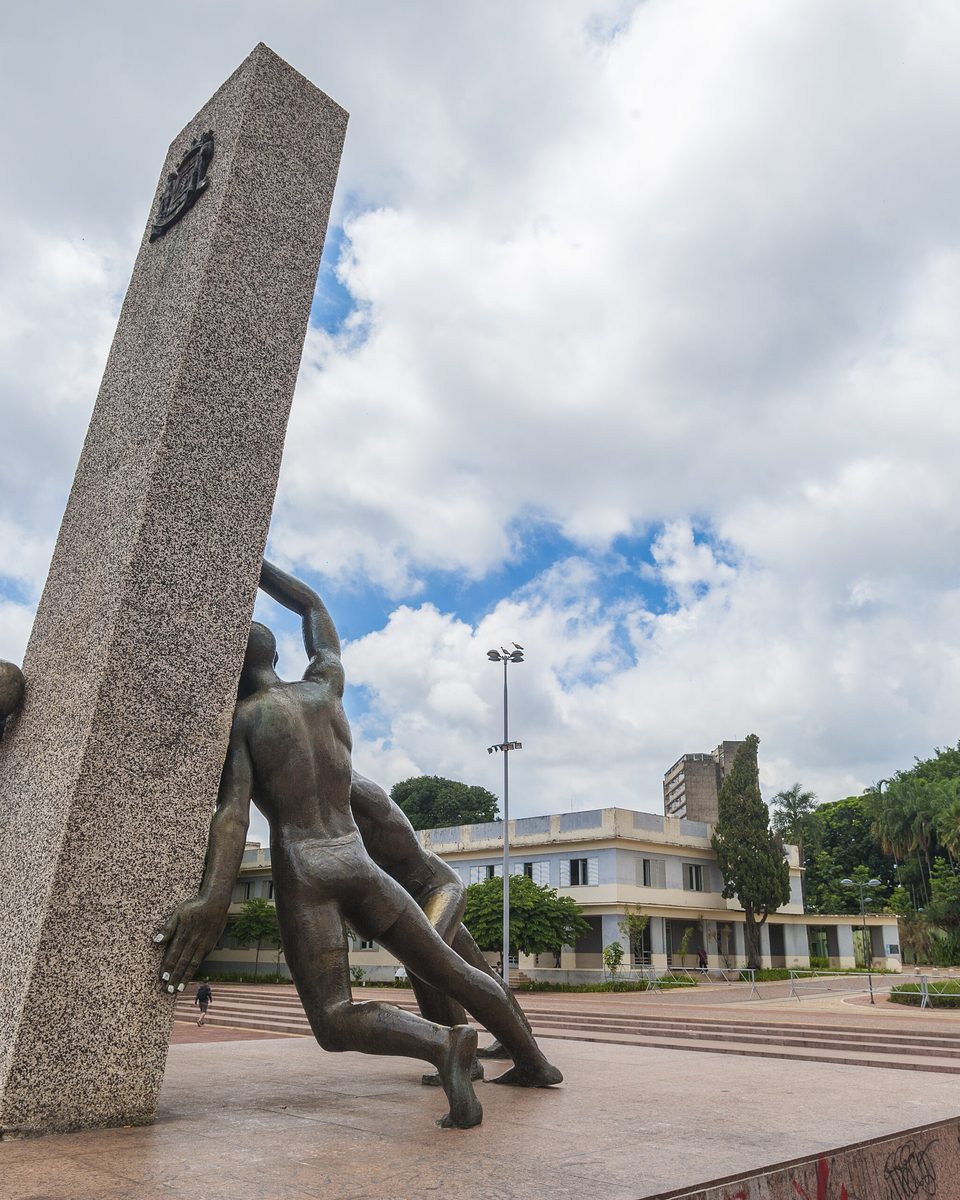 Monumento às Três Raças na Praça Cívica, em Goiânia, com três figuras de bronze empurrando o obelisco de granito, prédios públicos e bandeiras ao fundo, sob céu com nuvens.
