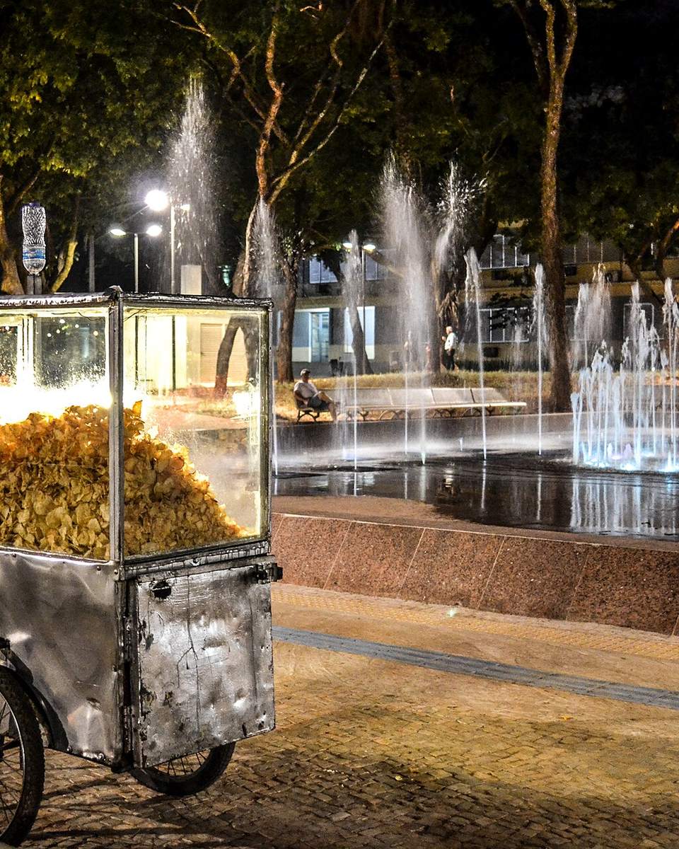 Carrinho de pipoca iluminado ao lado da Fonte Luminosa da Praça Cívica, em Goiânia, à noite, com os jatos d'água acesos, bancos e árvores ao fundo.