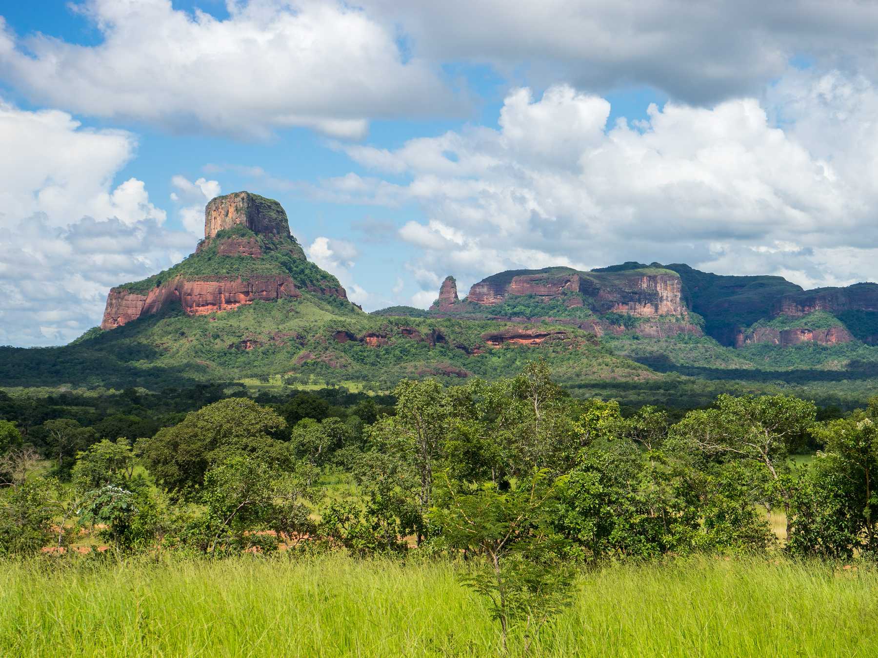 Morro de paredões avermelhados no alto da Serra dos Caiapós, na região do Pinga Fogo, com o cerrado verde em primeiro plano e nuvens brancas no céu.