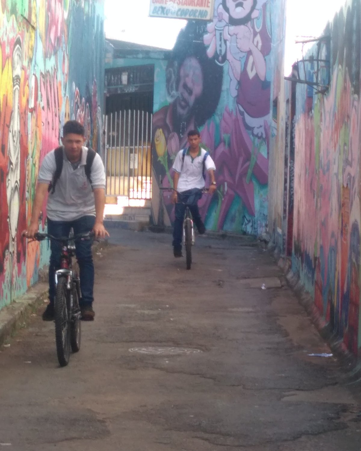 Duas pessoas de bicicleta num beco estreito do Centro de Goiânia com as paredes dos dois lados cobertas de grafites coloridos.