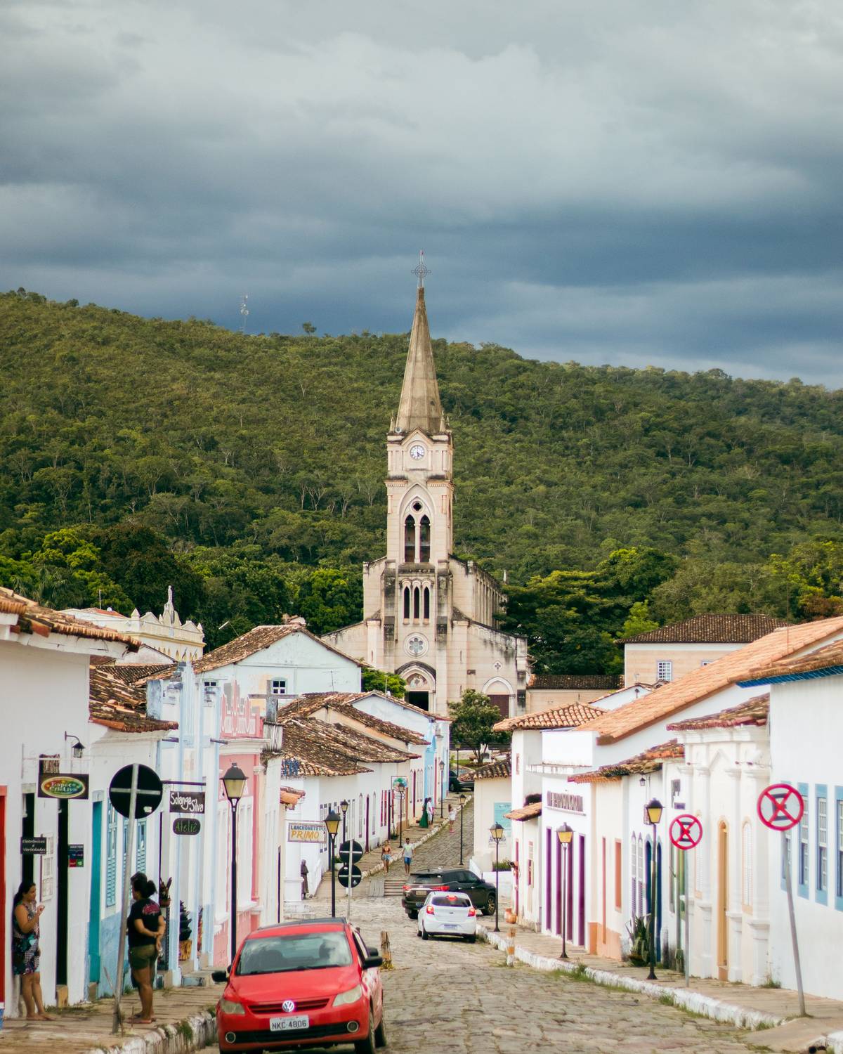 Rua de pedra da Cidade de Goiás com casas coloniais coloridas dos dois lados, um carro vermelho estacionado e a torre da Igreja do Rosário ao fundo, diante do morro verde.