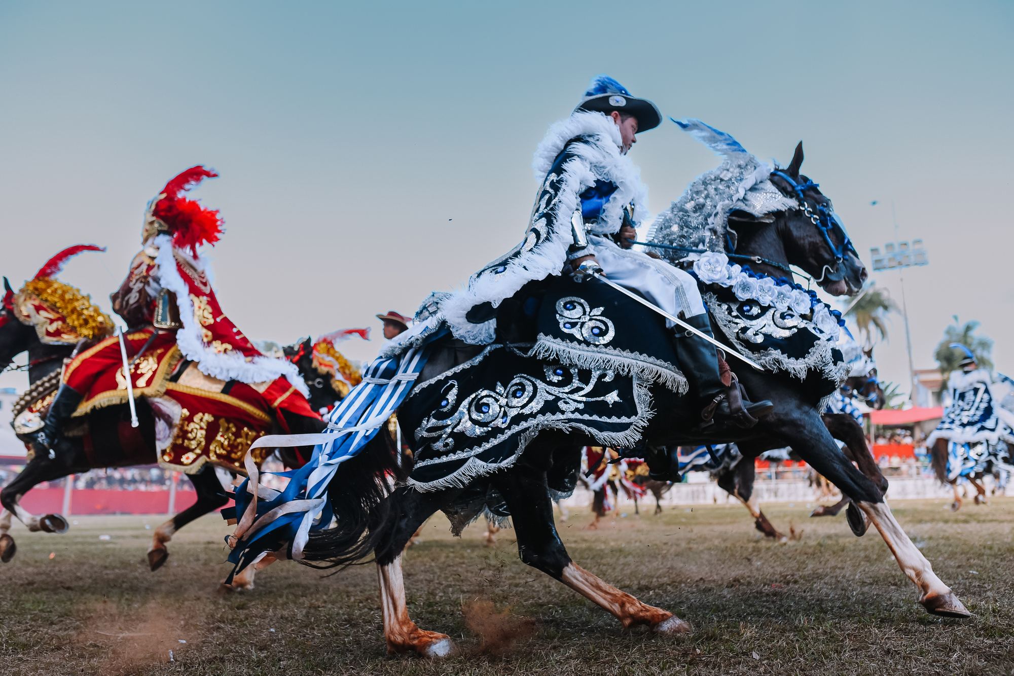 Cavaleiros das Cavalhadas de Pirenópolis galopando no campo, um deles num cavalo preto coberto de tecido bordado em preto e prata e de roupa branca com chapéu de plumas, e outro de vermelho e dourado ao lado.