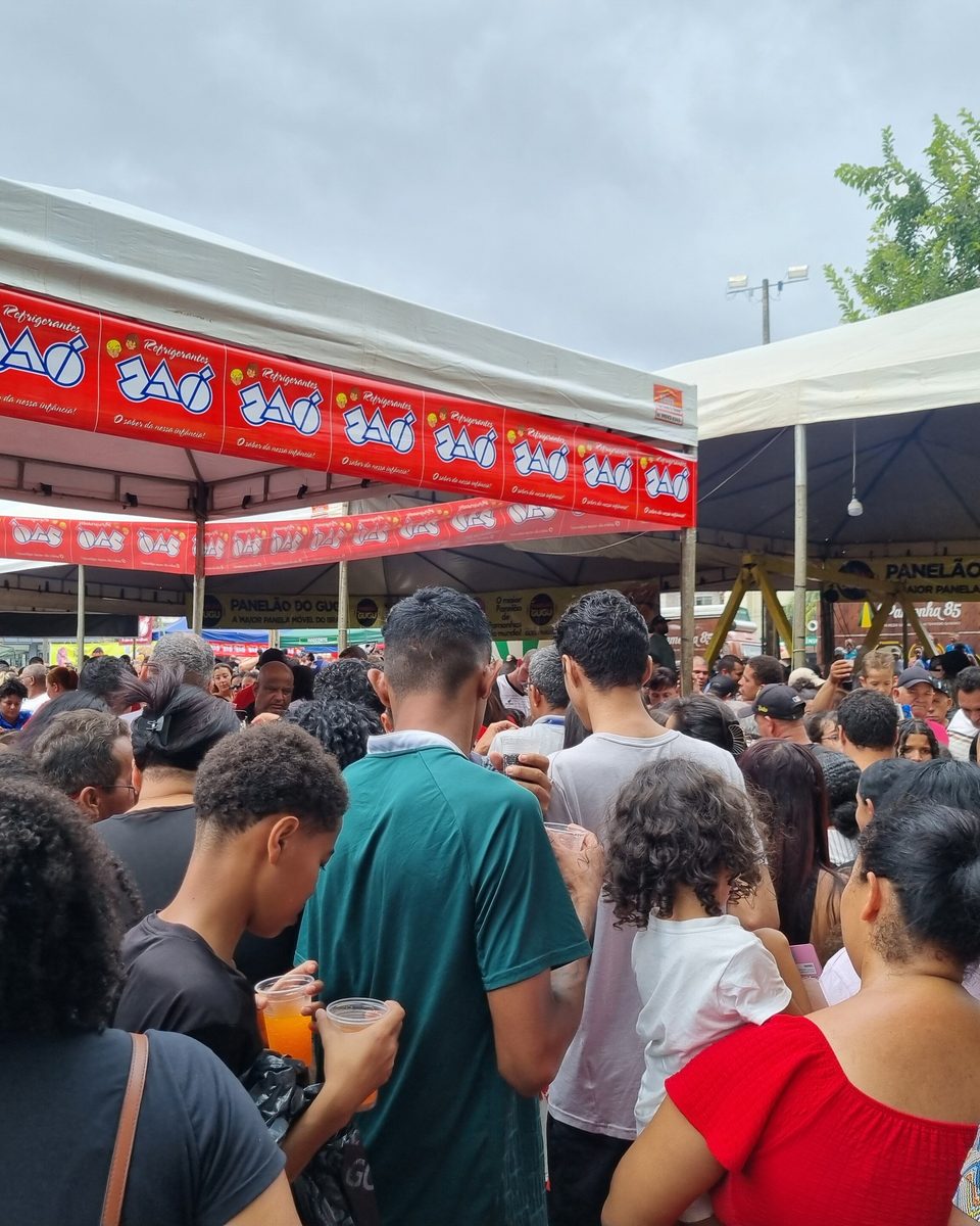 Muita gente reunida sob tendas de faixa vermelha num festival de pamonha na Praça do Violeiro, em Goiânia, com pessoas em pé conversando e esperando a vez.