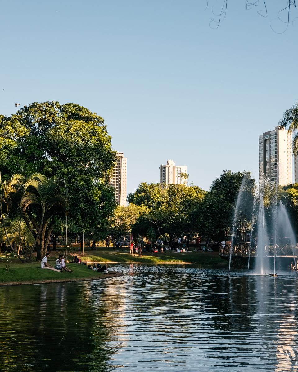Lago do Parque Vaca Brava, em Goiânia, com o chafariz jorrando, pessoas sentadas no gramado à sombra das árvores e prédios altos ao fundo.