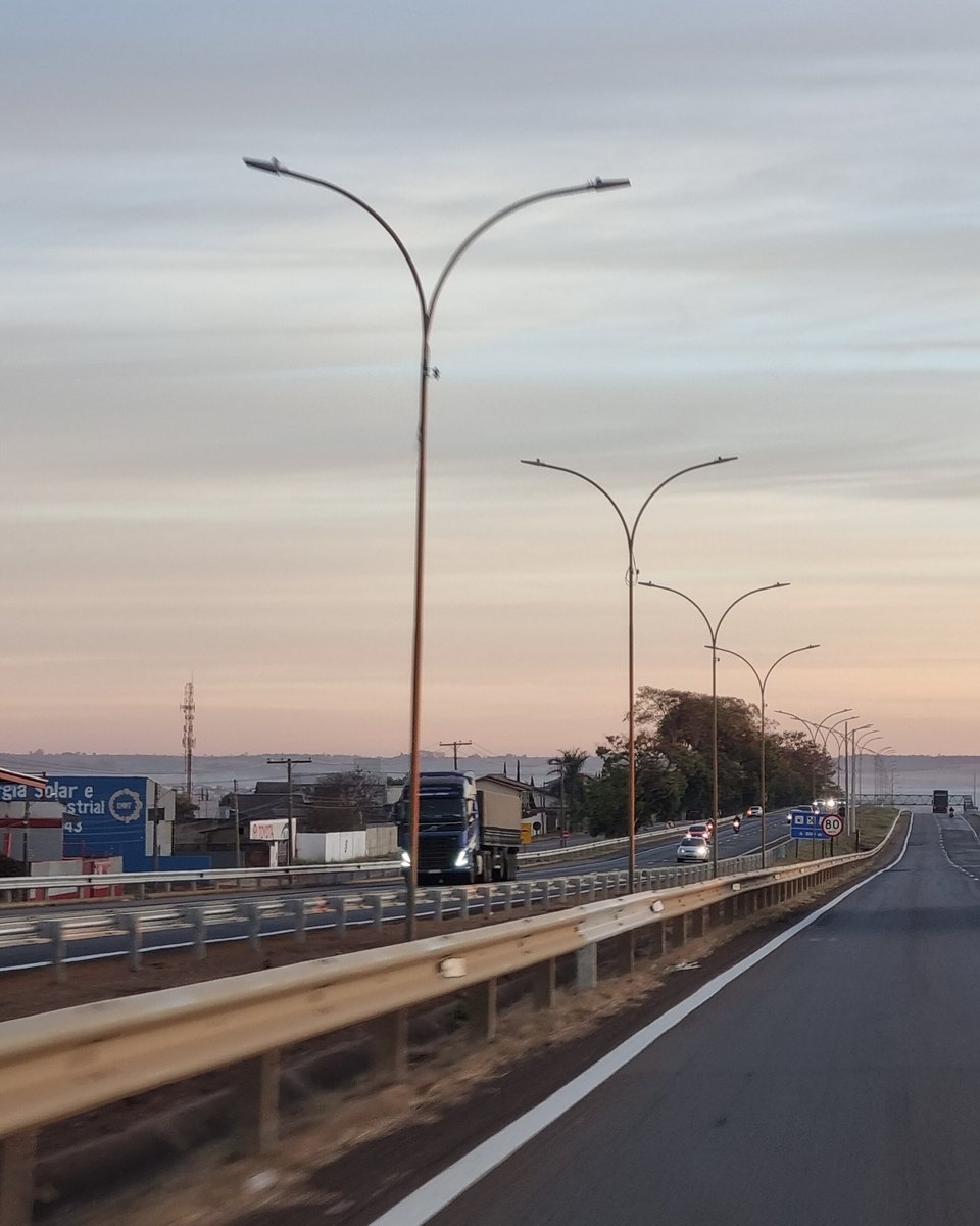 BR-153 em Anápolis com pista dupla, caminhões e carros trafegando, postes altos no canteiro central e o céu do fim de tarde.