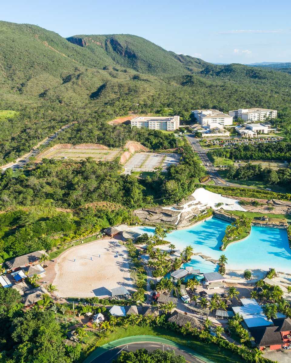 Vista aérea de um parque de águas quentes em Rio Quente, com piscinas de água verde-clara entre coqueiros e mata, quiosques de palha e a serra coberta de cerrado ao fundo.