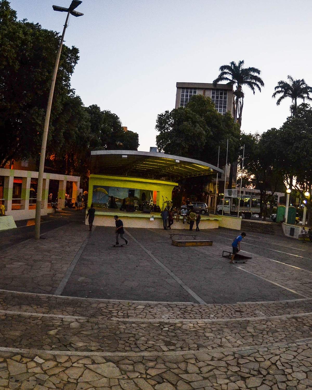 Praça dos Pioneiros, no Centro de Governador Valadares, ao entardecer, com gente na praça e postes acesos.