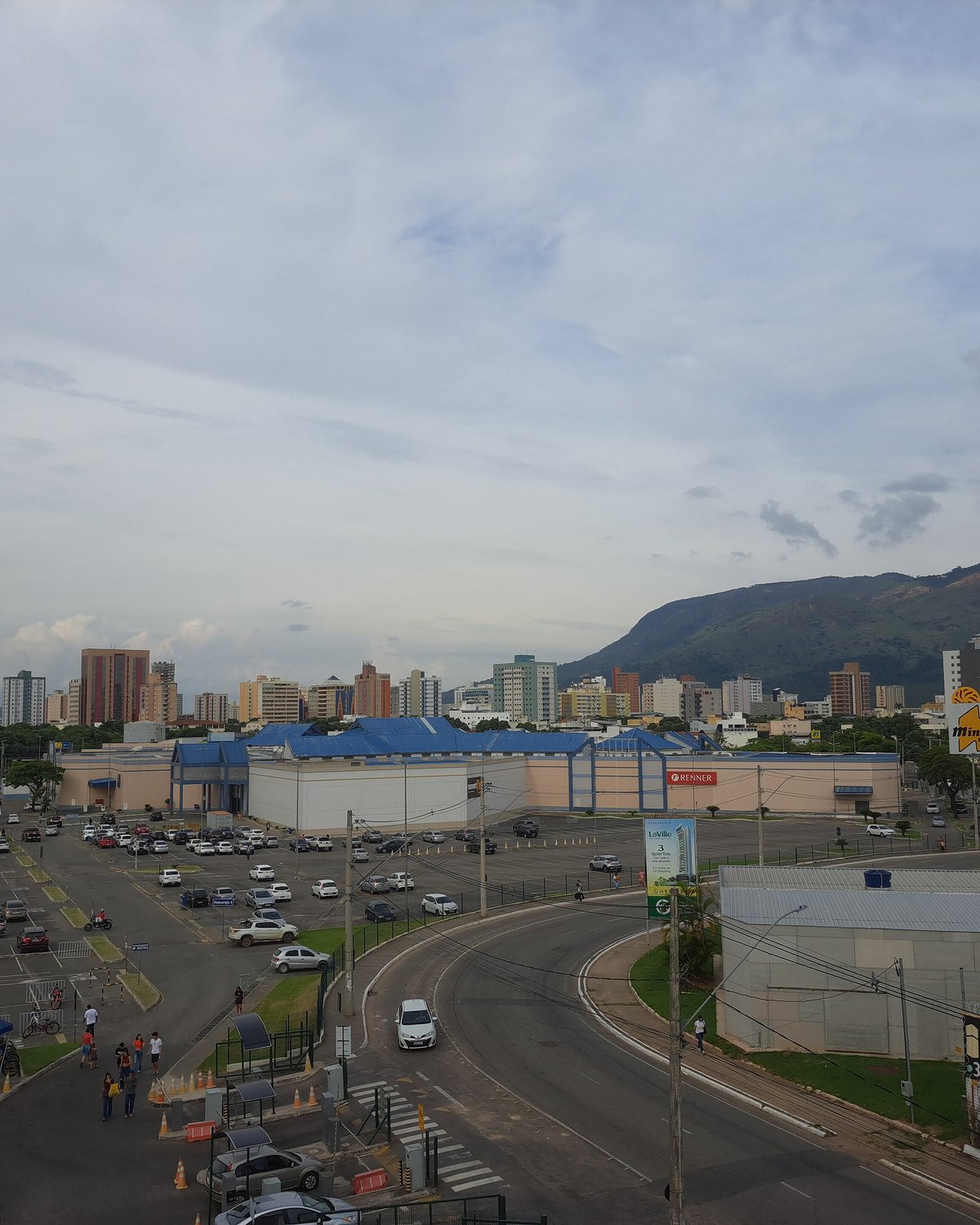 Vista do alto do Centro de Governador Valadares, com uma avenida larga, carros, galpões de telhado azul, prédios residenciais e o Pico da Ibituruna ao fundo sob um céu nublado.