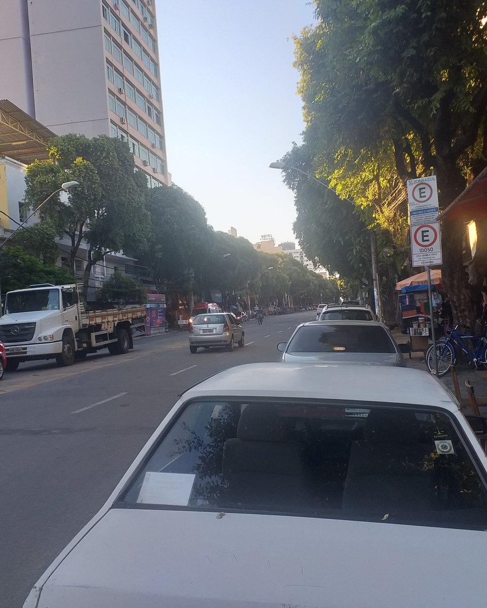 Trecho da Avenida Minas Gerais, no Centro de Governador Valadares, num fim de tarde, com lojas e toldo vermelho à direita, mesas de madeira na calçada, um homem de mochila caminhando, carros e um caminhão estacionados e prédios altos ao fundo.