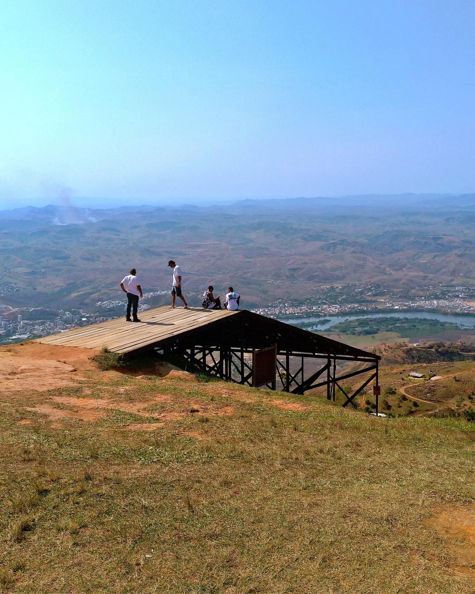 Rampa de madeira de decolagem de voo livre no alto do Pico da Ibituruna, com algumas pessoas de pé e sentadas na ponta, o gramado seco em primeiro plano e, lá embaixo, a cidade de Governador Valadares e o Rio Doce sob a névoa azulada.