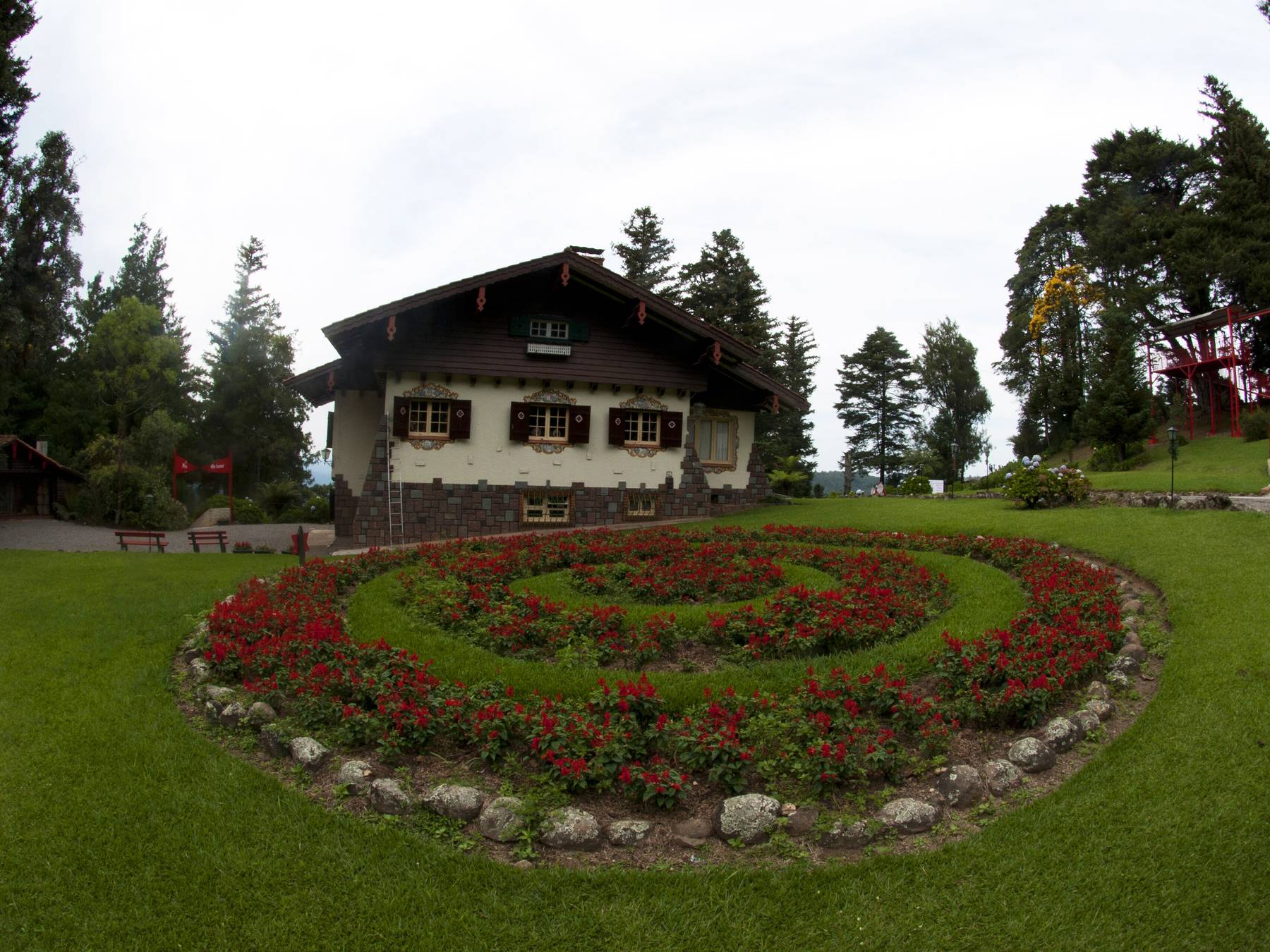 Jardim com canteiro circular de flores vermelhas no gramado da Aldeia do Papai Noel, com uma casa em estilo alpino e pinheiros ao fundo.