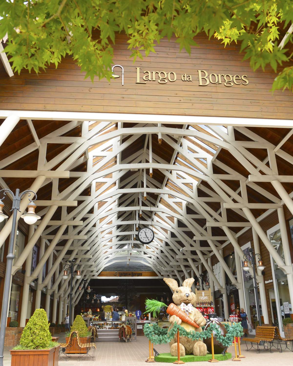 Galeria coberta do Largo da Borges, em Gramado, com estrutura de madeira branca, lojas dos dois lados, vasos de plantas e decoração de Páscoa no corredor.