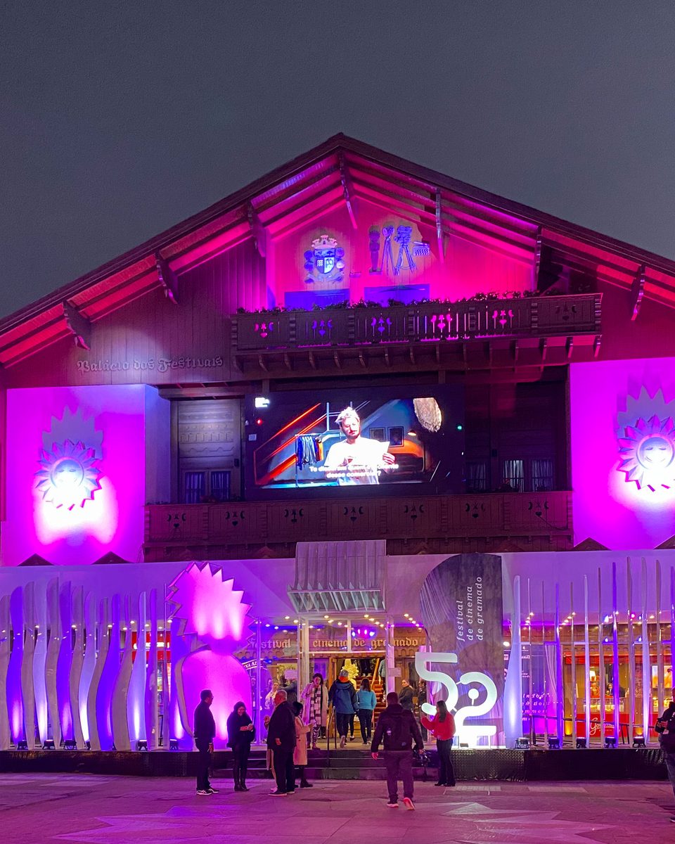 Palácio dos Festivais, em Gramado, iluminado de rosa e roxo à noite durante o Festival de Cinema, com público na calçada.