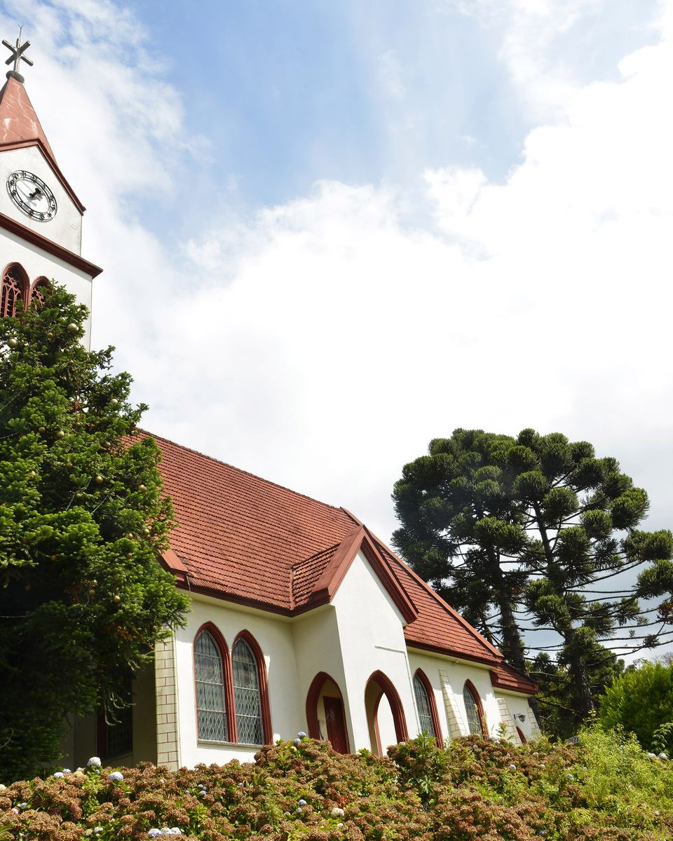 Igreja do Relógio, em Gramado, branca, de telhado vermelho e torre com relógio, cercada de araucárias e canteiros de flores, sob céu azul com nuvens.