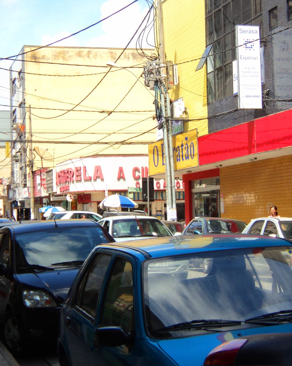 Rua de comércio no Centro de Ipatinga com lojas, carros estacionados e pedestres na calçada.