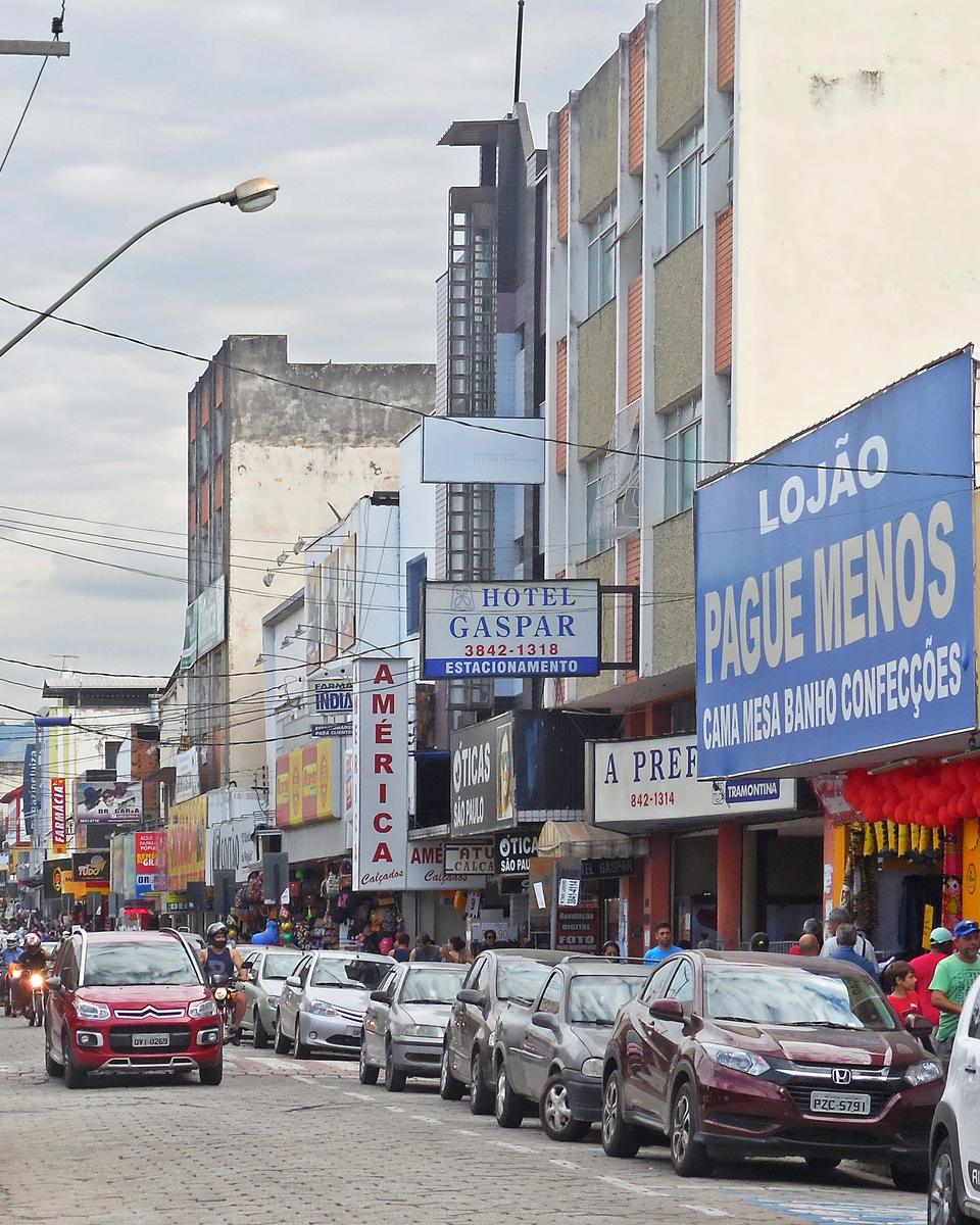 Rua Pedro Nolasco, no centro de Coronel Fabriciano, com lojas de fachadas coloridas dos dois lados, placas de lanchonete e de loja de cama, mesa e banho, carros estacionados e pessoas nas calçadas num dia nublado.