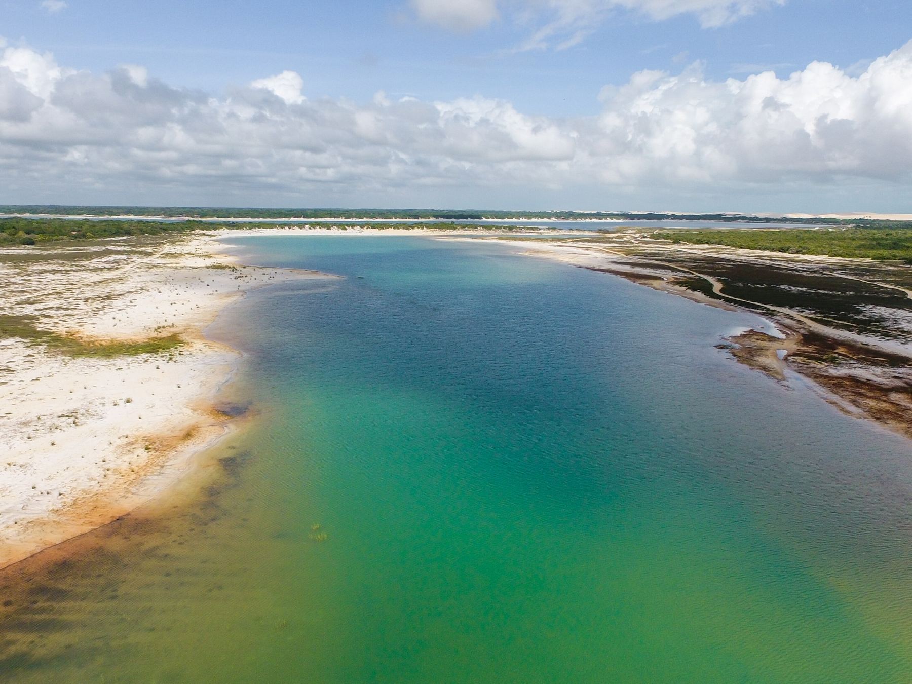 Vista aérea da Lagoa Azul, uma faixa longa de água verde e azul-turquesa entre margens de areia branca e vegetação rasteira, com as dunas e o céu de nuvens brancas no horizonte.