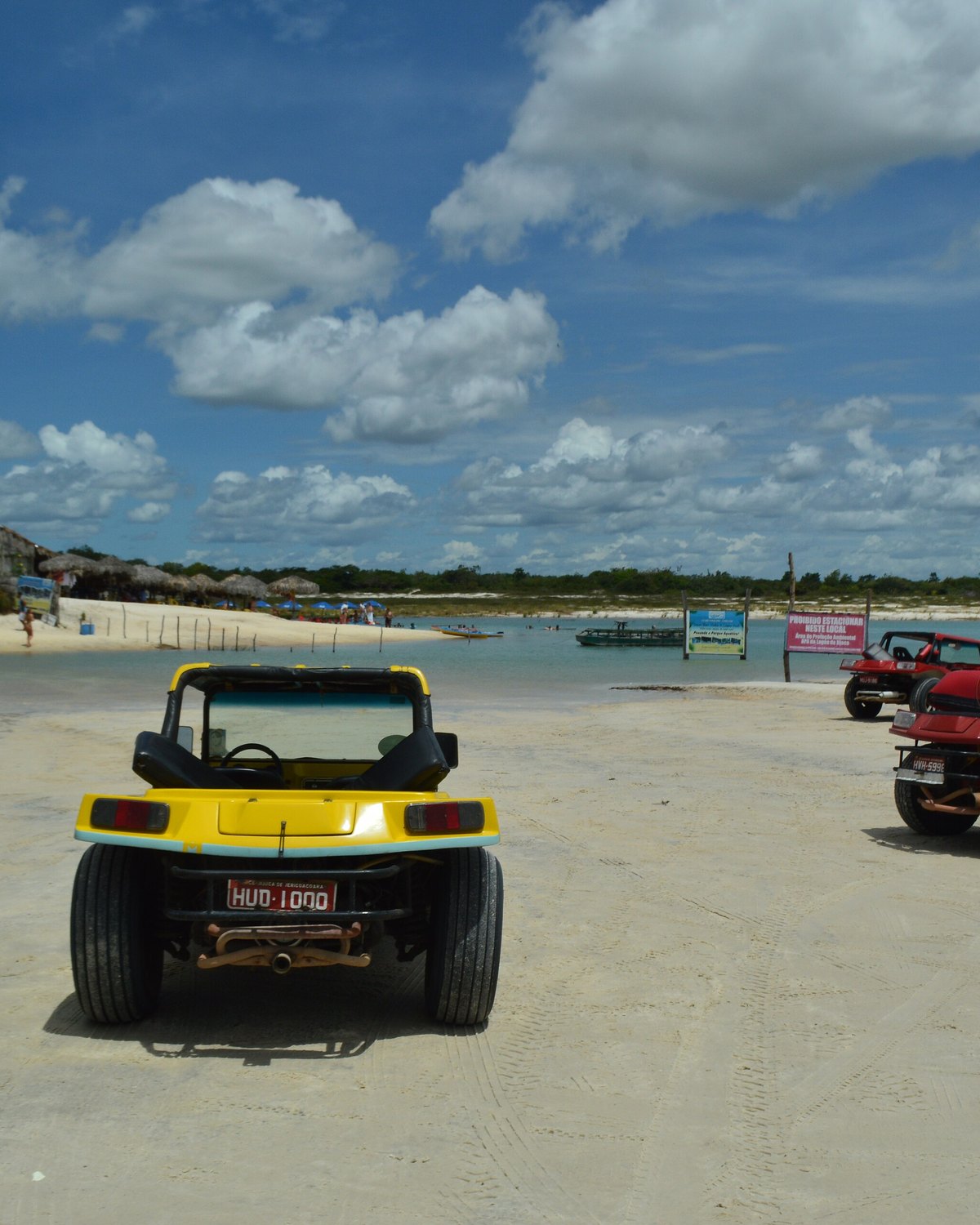 Buggys amarelos e vermelhos estacionados na areia da Lagoa Azul, com a água, barracas e outros buggys ao fundo sob céu azul com nuvens.