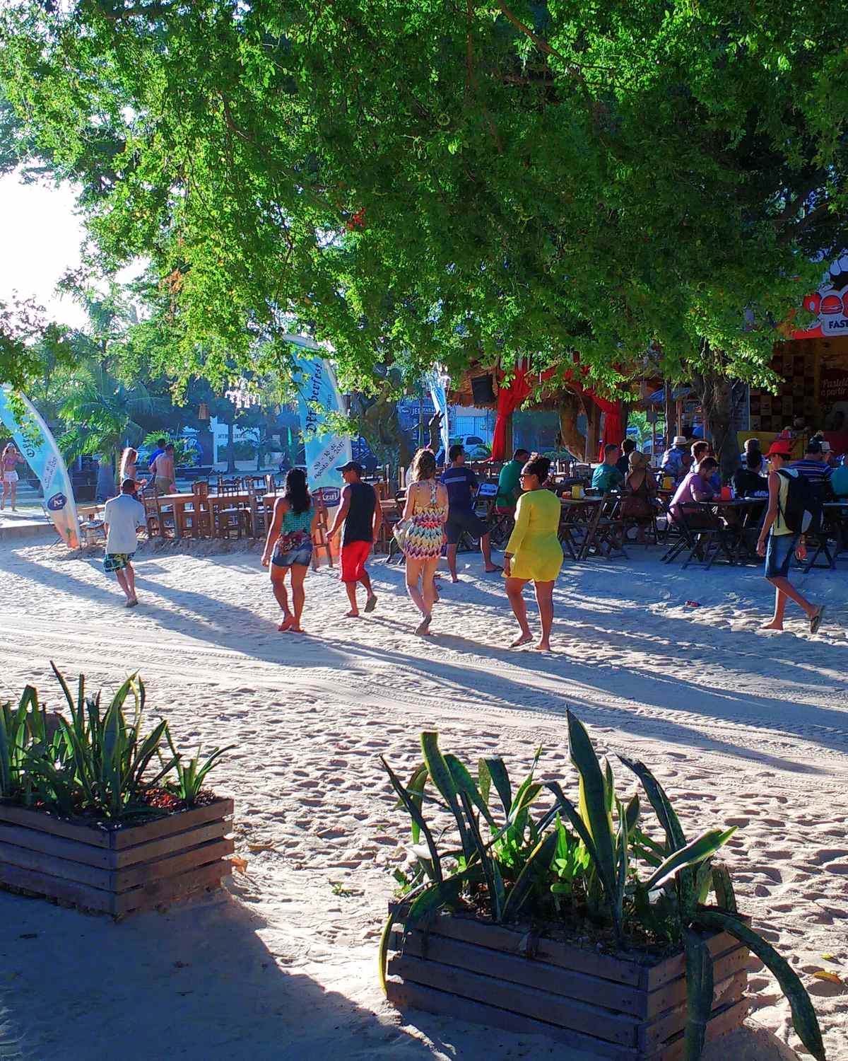 Turistas caminhando pela areia da vila de Jericoacoara, à sombra de árvores.