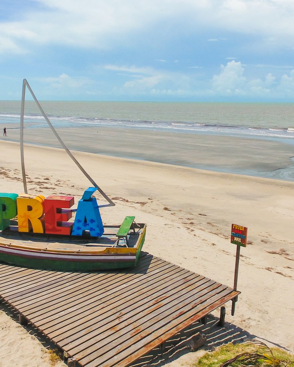 Letreiro colorido com a palavra Preá sobre um deck de madeira na areia, ao lado de um barco com vela, com a praia larga e o mar ao fundo.