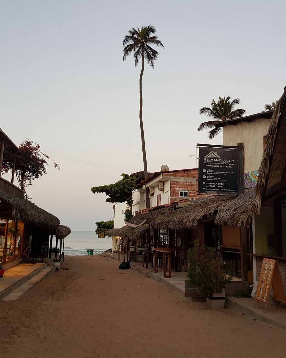 Rua de areia da vila de Jericoacoara ao entardecer, com lojas e restaurantes de telhado de palha dos dois lados, um coqueiro alto e o mar no fim da rua.
