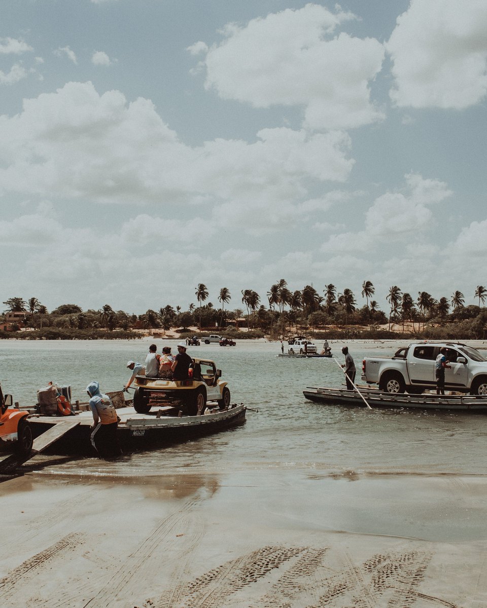 Buggy laranja e caminhonete atravessando de balsa um braço de rio raso, com balseiros empurrando as balsas com varas e coqueiros na outra margem.