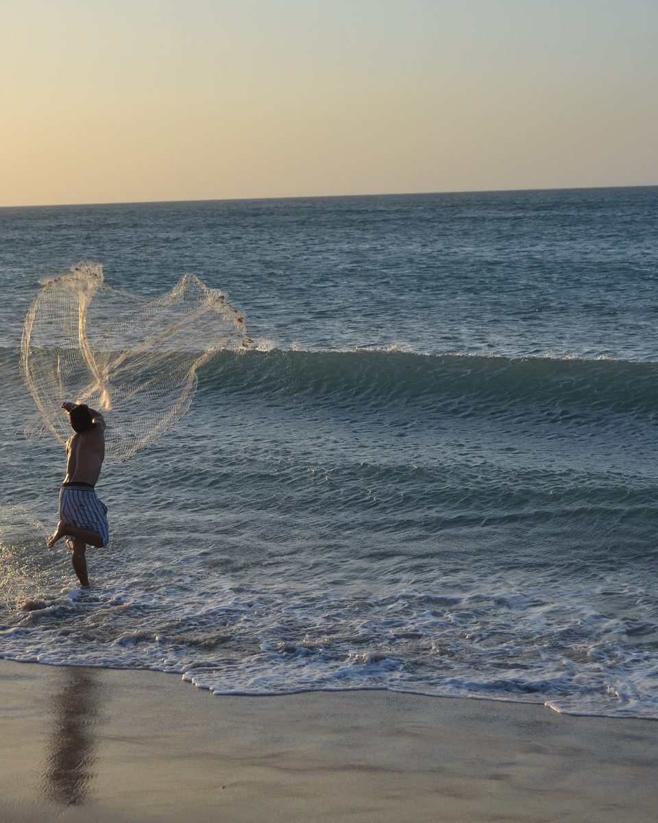 Pescador lançando a tarrafa aberta no ar sobre as ondas da praia de Jericoacoara, no fim da tarde.
