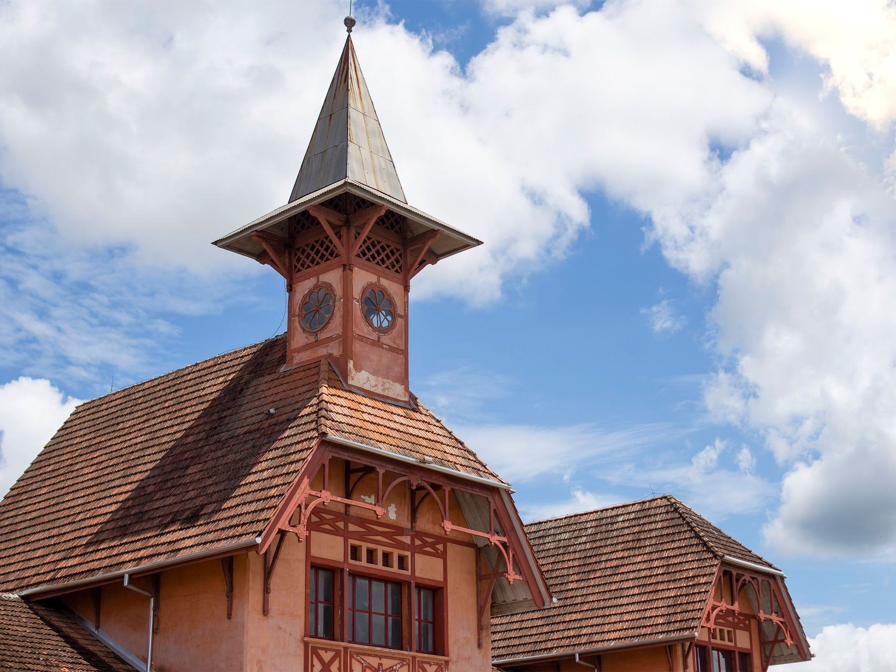 Torre com relógio e telhado pontiagudo da Estação da Memória, antiga estação ferroviária de Joinville, com detalhes de madeira na fachada cor de tijolo, contra o céu azul com nuvens brancas.