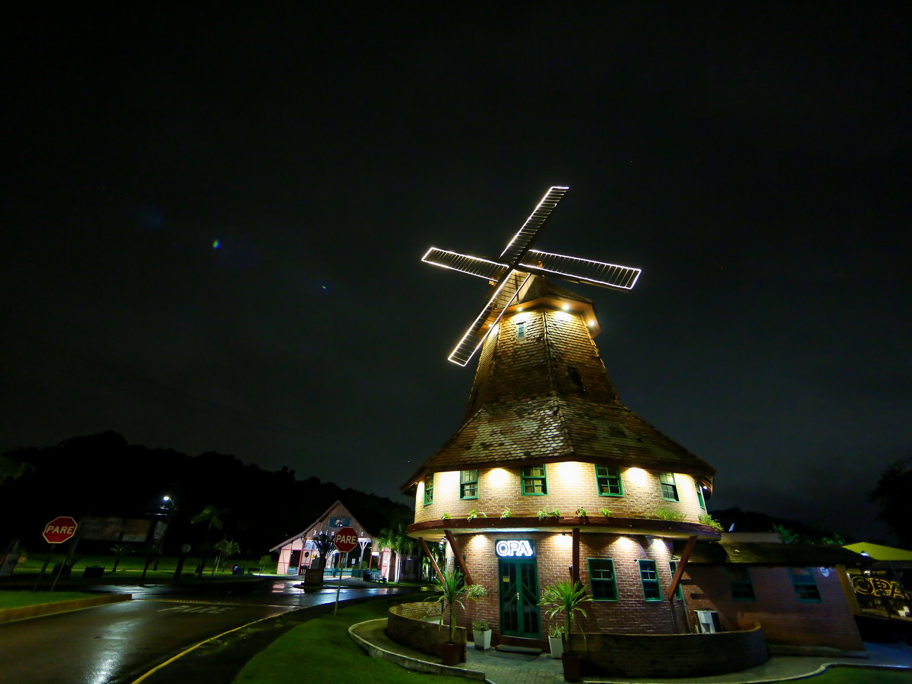 Moinho de vento com pás iluminadas por luzes brancas na entrada de Joinville, à noite, com a base de pedra e janelas acesas, gramado em volta e a avenida molhada de chuva.