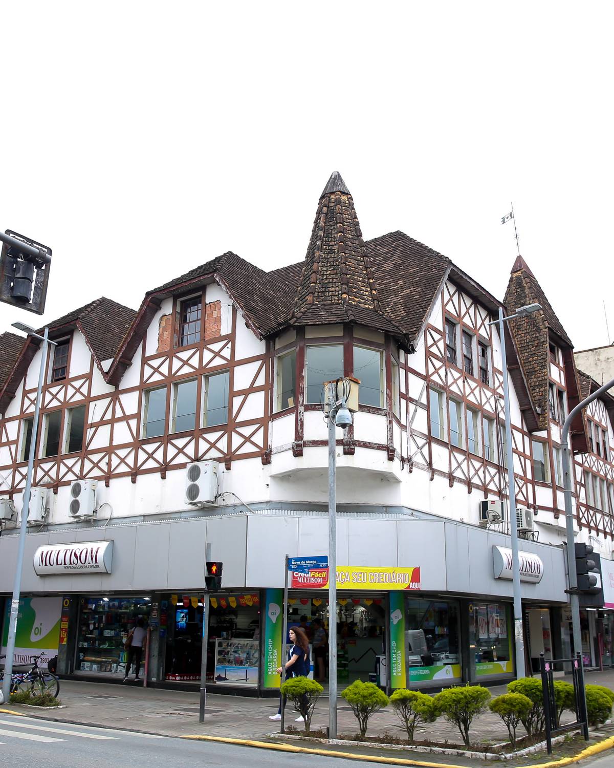 Prédio comercial em estilo enxaimel, com vigas escuras aparentes na fachada branca, torres pontiagudas e lojas no térreo, numa esquina com semáforos no Centro de Joinville.