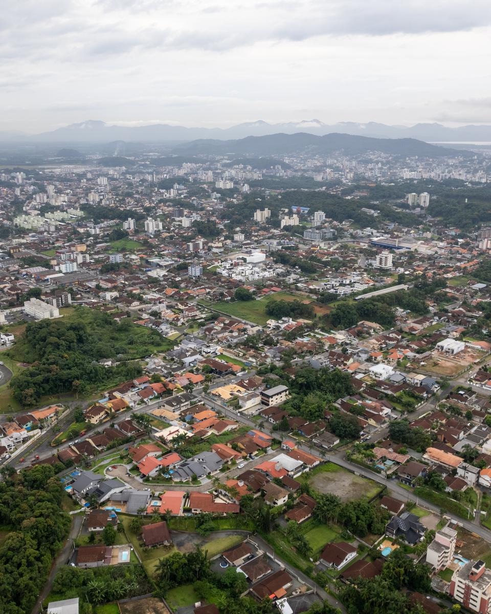 Vista aérea de Joinville num dia nublado, com casas de telhado vermelho, prédios e ruas arborizadas até os morros verdes da Serra do Mar no horizonte.