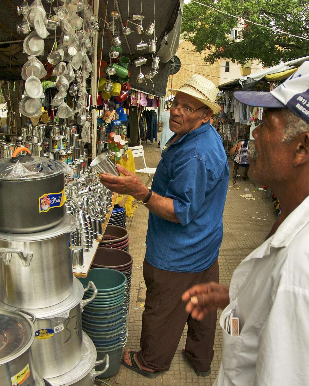 Dois senhores, um de chapéu de palha e outro de boné, olhando panelas de alumínio, baldes e utensílios pendurados numa barraca de rua em Juazeiro do Norte.