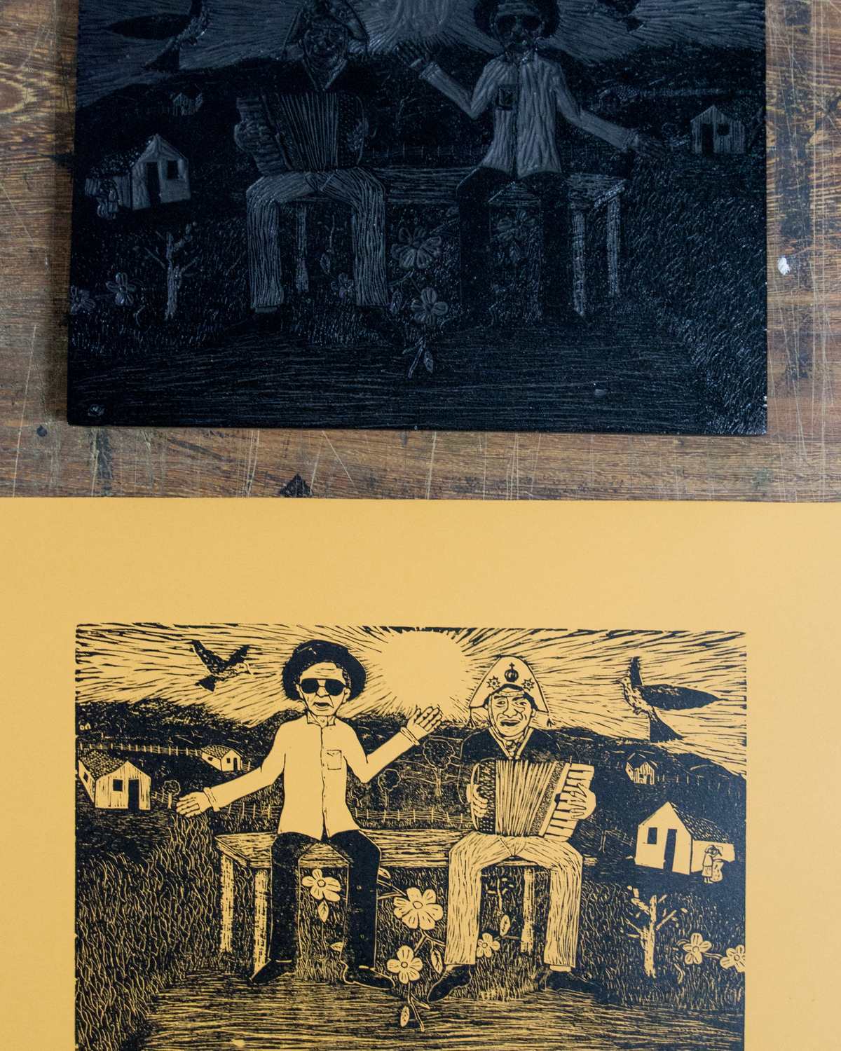Matriz de xilogravura entalhada em madeira escura e, embaixo, a gravura impressa em papel amarelo, com um sanfoneiro e um homem de chapéu diante de casinhas do sertão e pássaros no céu.