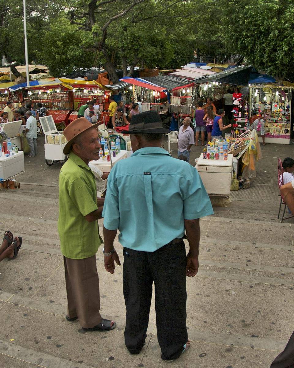 Senhores de chapéu conversando no alto de uma escadaria, em Juazeiro do Norte, diante de um largo cheio de barracas coloridas com toldos e gente circulando entre elas.