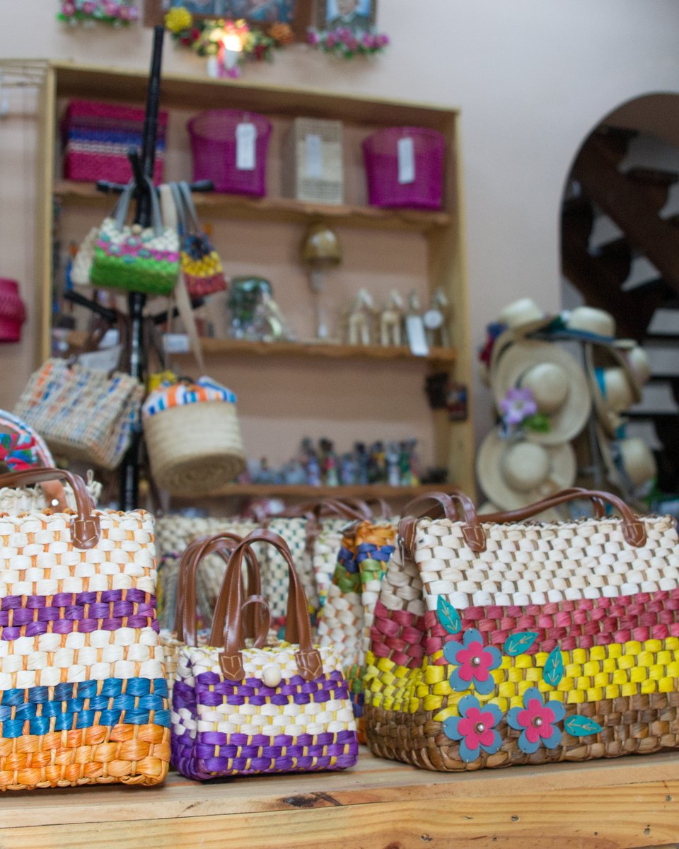 Bolsas de palha trançada com listras roxas, amarelas, laranja e brancas e flores bordadas, expostas sobre uma mesa numa loja de artesanato, com prateleiras de bolsas, chapéus e lembranças ao fundo.
