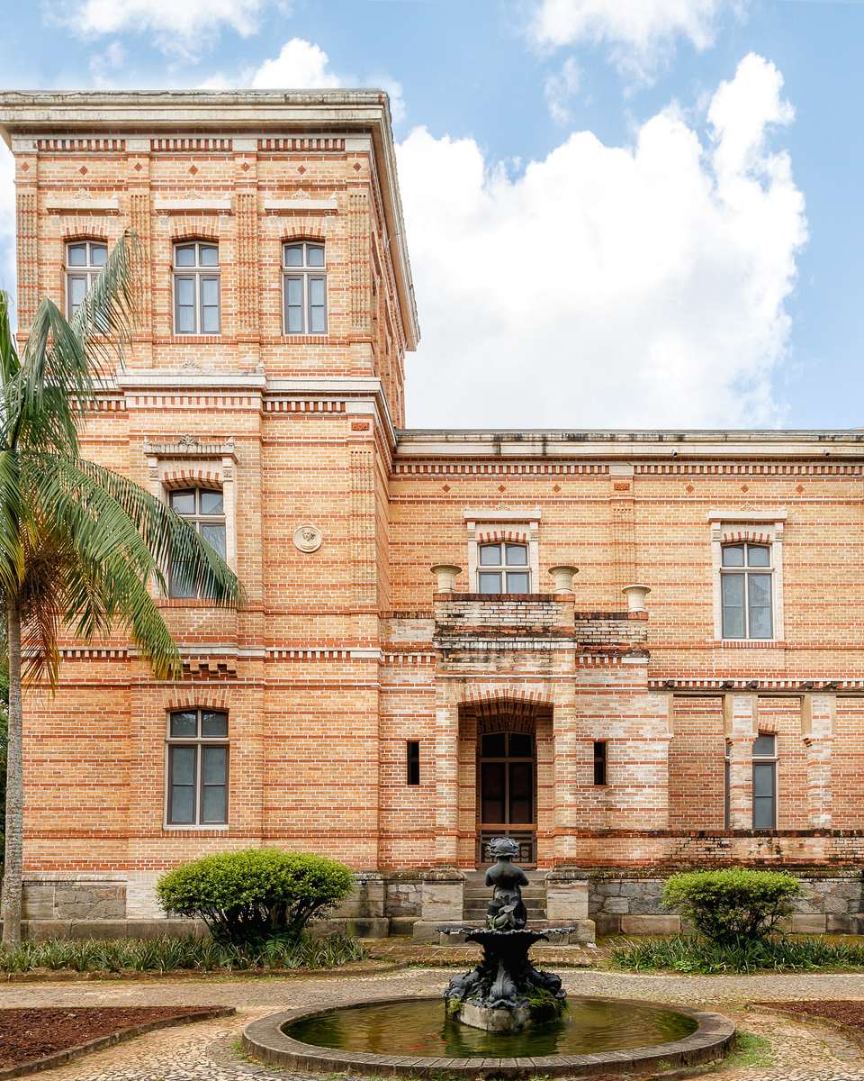 Prédio de tijolos aparentes do Museu Mariano Procópio, em Juiz de Fora, com palmeira e chafariz na frente.
