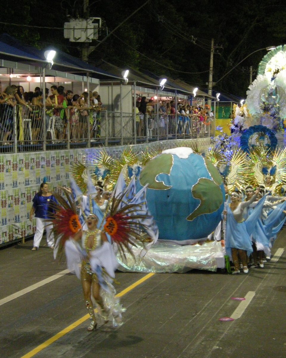 Comissão de frente da escola de samba Unidos do Ladeira desfilando à noite no carnaval de Juiz de Fora, com passista de fantasia branca e plumas, carro alegórico em forma de globo e o público na arquibancada.